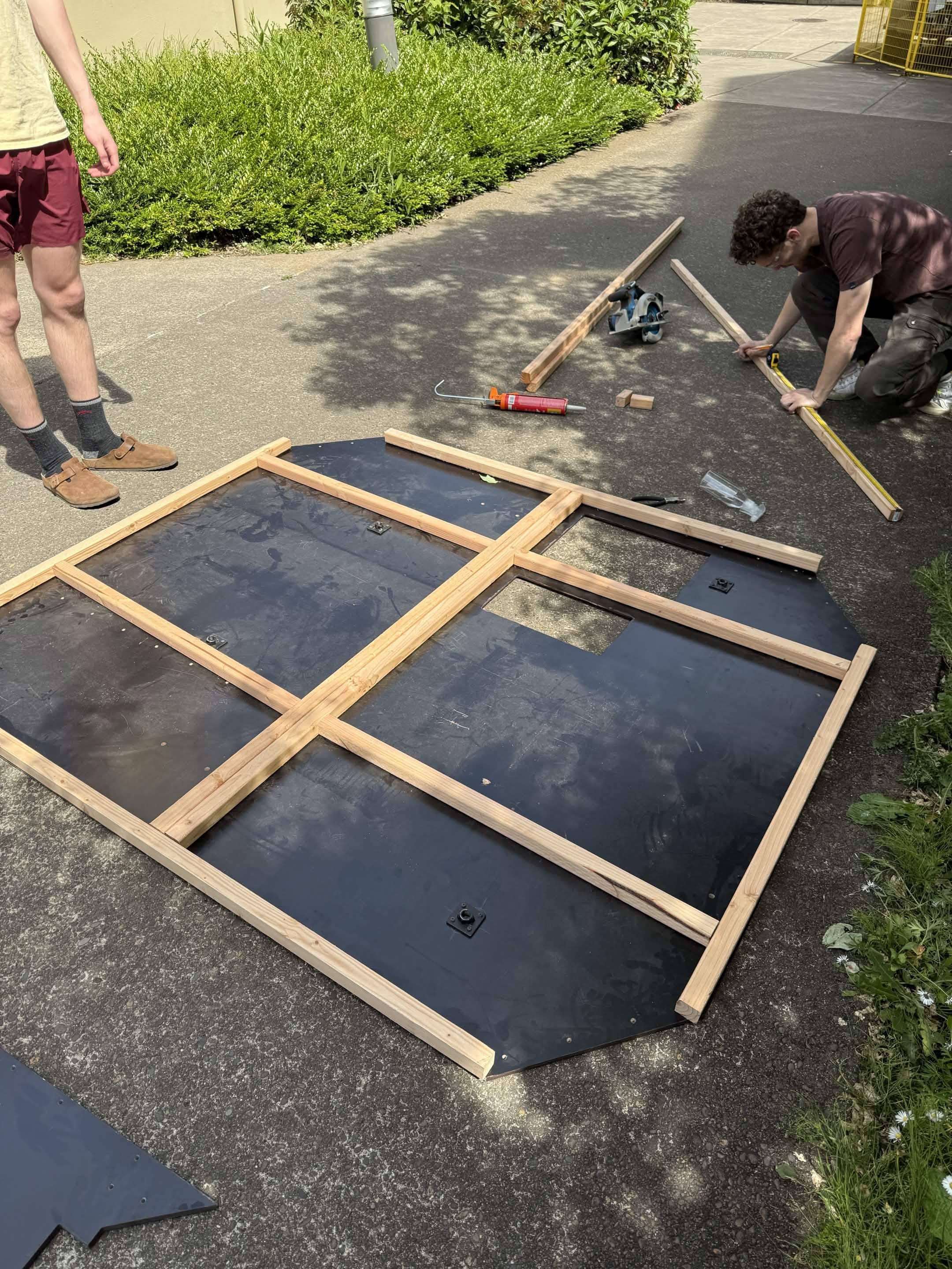 Building the raised platform: a 2×2 lumber support frame fastened to the underside of the black platform panels, with teammates measuring and cutting outside.